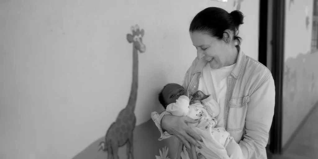 Debra Mace smiles while holding a baby in her arms, standing next to a wall with a giraffe illustration.